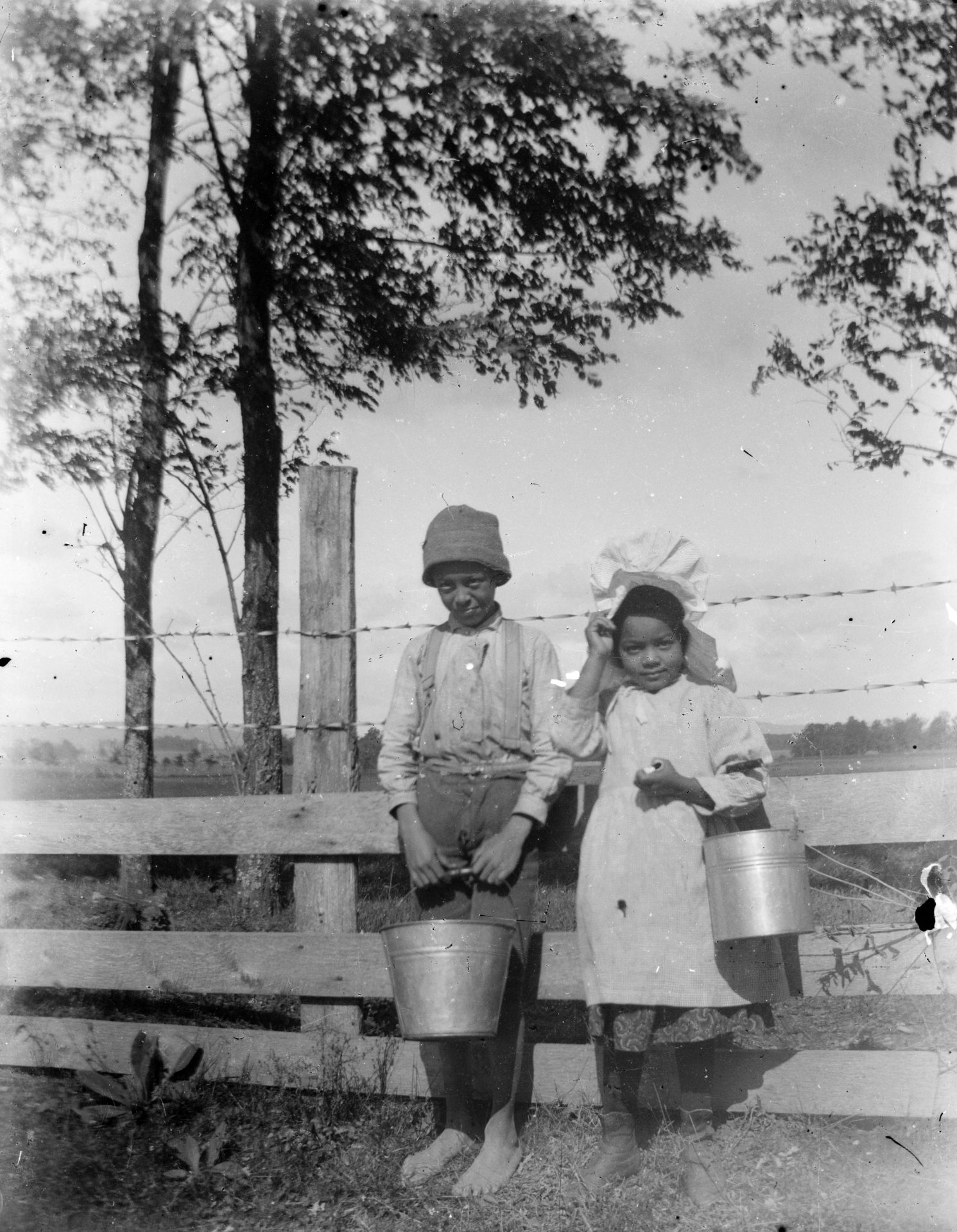 An African American boy and girl hold milk pails and pose in front of a fence.